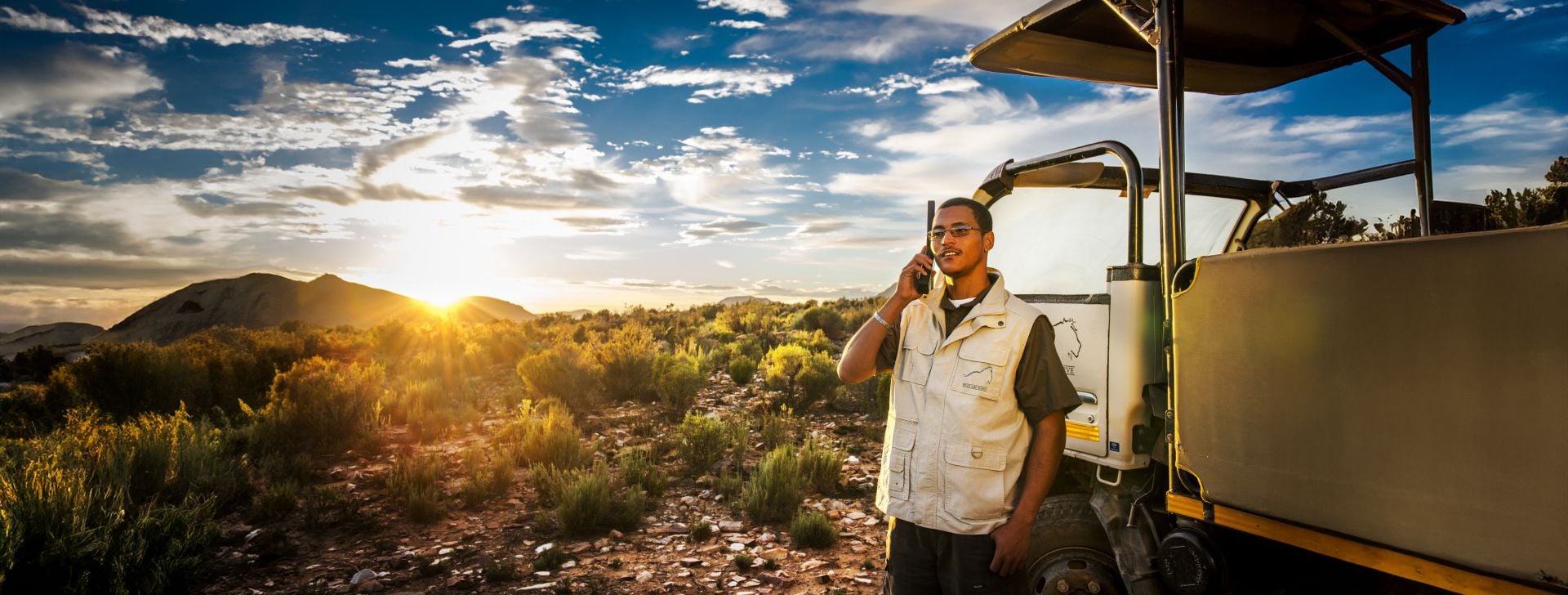 Field researcher talking on a phone beside an off-road vehicle in a remote landscape at sunset.